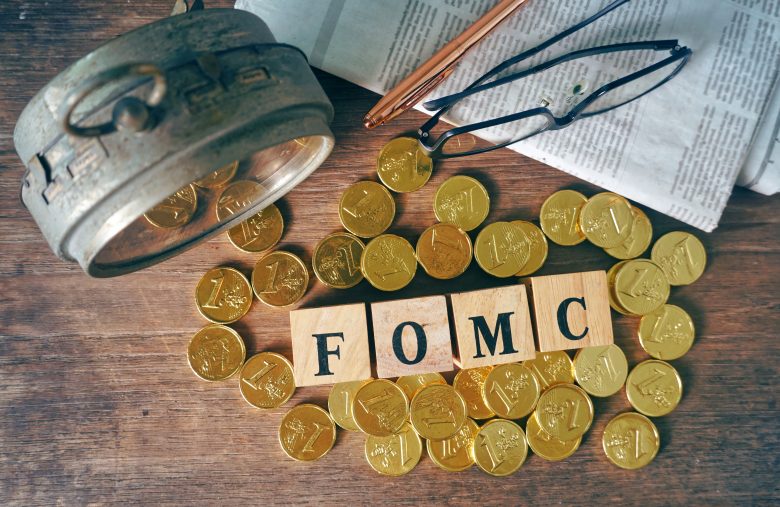 Top view wood cube which have text "FOMC" decorate with gold coins near old clock and pen and glasses on the newspaper,econimic data concept.